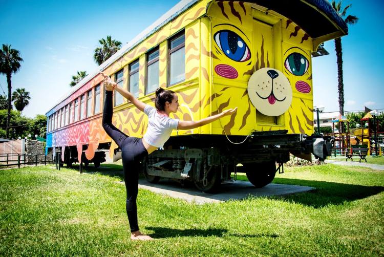 Sandra haciendo una postura de yoga en un parque, con un tren amarillo de fondo.